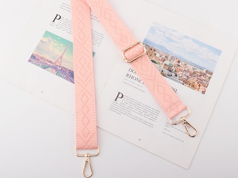 May include: A light pink bag strap with a geometric pattern and gold-colored metal hardware. The strap is lying on a magazine with images of a city and the Eiffel Tower.