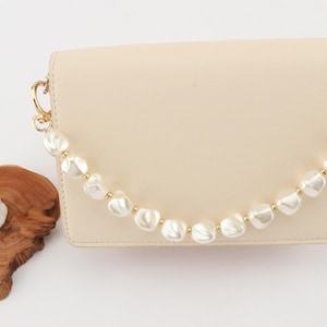 May include: A cream-colored handbag with a pearl beaded handle and gold-tone hardware. The bag is rectangular with a flap closure. The pearl handle is made of large, irregular-shaped white pearls and small gold beads.