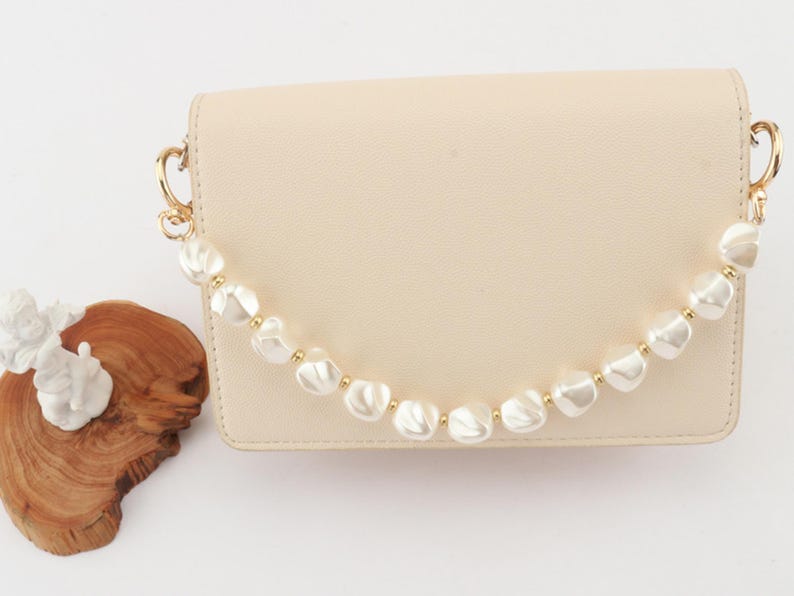 May include: A cream-colored handbag with a pearl beaded handle and gold-tone hardware. The bag is rectangular with a flap closure. The pearl handle is made of large, irregular-shaped white pearls and small gold beads.