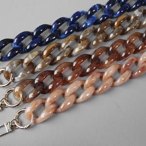 May include: Four acrylic chain straps for bags, each featuring large interlocking oval links with a marbled finish. The colours include deep blue, dark brown, medium brown, and light beige. Each strap is finished with silver-tone metal lobster clasps.