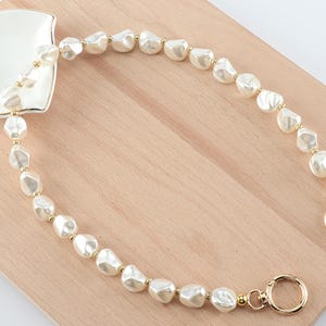 May include: A cream-colored pearl beaded bag strap with gold-colored hardware. The strap is made of irregular-shaped pearls and is attached to a small, square, white dish with a gold trim. The strap is on a light brown wooden surface.