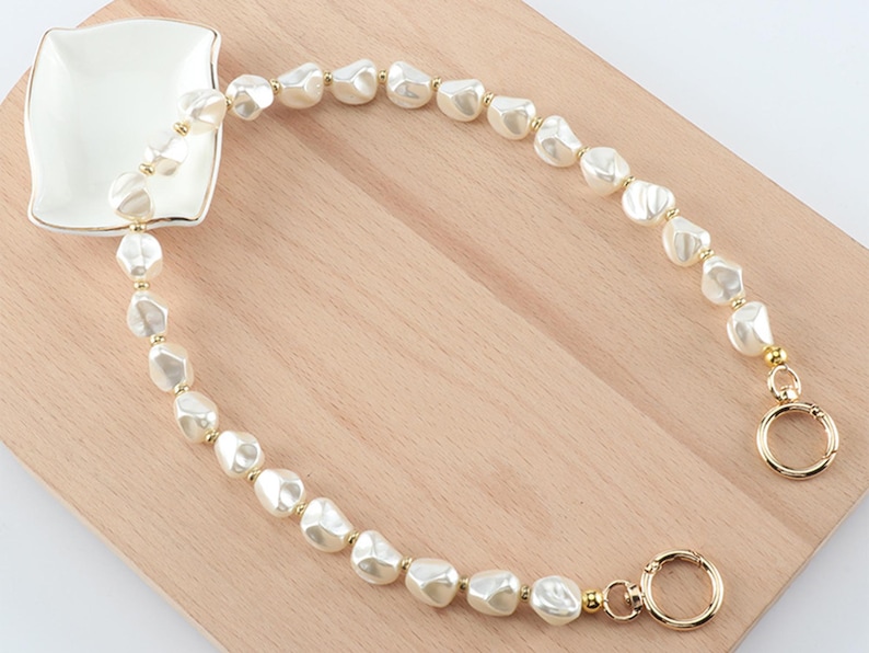 May include: A cream-colored pearl beaded bag strap with gold-colored hardware. The strap is made of irregular-shaped pearls and is attached to a small, square, white dish with a gold trim. The strap is on a light brown wooden surface.