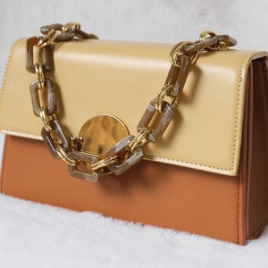 May include: A rectangular handbag featuring a two-tone design with a light beige top flap and a tan base. The bag includes a decorative chunky chain strap made of translucent brown marbled links and gold-toned metal, centered with a circular gold disc.