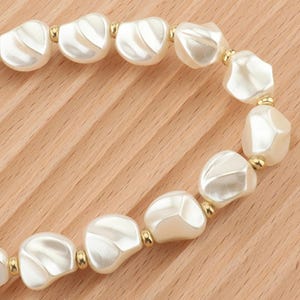 May include: A close-up of a necklace featuring large, irregular, off-white beads with a pearlescent sheen. The beads are connected by small gold-colored metal beads. The necklace is displayed on a light brown wooden surface.