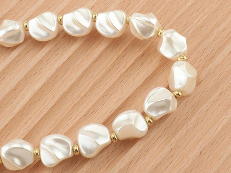 May include: A close-up of a necklace featuring large, irregular, off-white beads with a pearlescent sheen. The beads are connected by small gold-colored metal beads. The necklace is displayed on a light brown wooden surface.