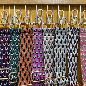May include: Nine colorful patterned fabric straps with gold metal clasps hanging on hooks. The straps are all different colors and patterns, including red, green, orange, purple, brown, and pink.