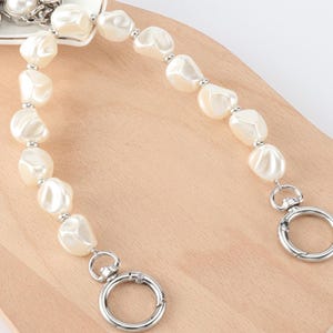 May include: A cream-colored pearl beaded purse strap with silver-tone metal clasps. The strap features large, irregular-shaped pearls strung together with small silver beads. The clasps are round and open with a lever.