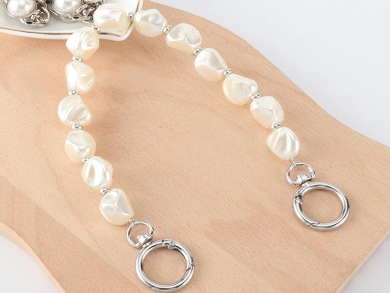 May include: A cream-colored pearl beaded purse strap with silver-tone metal clasps. The strap features large, irregular-shaped pearls strung together with small silver beads. The clasps are round and open with a lever.