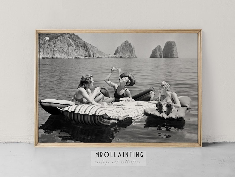 Peut inclure: Trois femmes en maillots de bain vintage se d&eacute;tendent sur un radeau gonflable ray&eacute; dans la mer. Elles profitent d'un pique-nique avec des boissons et des en-cas. L'arri&egrave;re-plan pr&eacute;sente des &icirc;les rocheuses au loin.