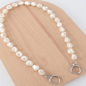 May include: A cream-colored pearl handbag strap with silver-tone metal hardware. The strap is composed of irregularly shaped pearls strung together with small silver beads. The strap has two circular clasps for attachment.
