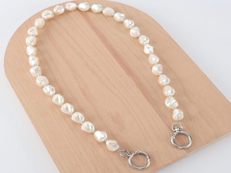 May include: A cream-colored pearl handbag strap with silver-tone metal hardware. The strap is composed of irregularly shaped pearls strung together with small silver beads. The strap has two circular clasps for attachment.