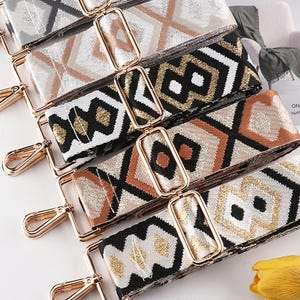 May include: Close-up of several patterned bag straps with gold-colored hardware. The straps feature geometric designs in various colors, including black, white, gold, and brown. The straps are likely for handbags or purses.