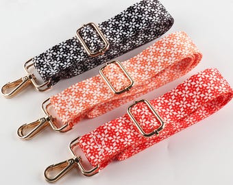 Könnte beinhalten: Drei gemusterte Taschenriemen in Schwarz, Orange und Rot, jeweils mit einem weißen Blumenmuster. Die Riemen verfügen über goldfarbene Metallbeschläge, darunter verstellbare Schnallen und Drehverschlüsse.