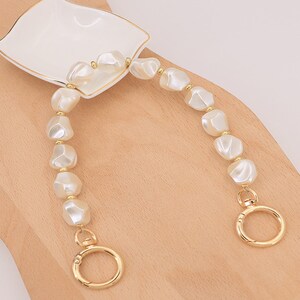 May include: A pearl-like beaded handbag strap with gold-colored hardware. The strap features large, irregular-shaped white beads and gold-toned connectors. The strap is attached to a white dish with a gold rim.
