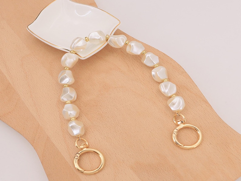 May include: A pearl-like beaded handbag strap with gold-colored hardware. The strap features large, irregular-shaped white beads and gold-toned connectors. The strap is attached to a white dish with a gold rim.