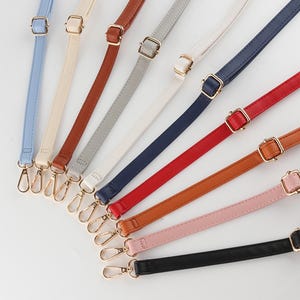 May include: A collection of colorful purse straps in various shades, including blue, cream, brown, gray, white, navy, red, orange, pink, and black. Each strap features a gold-colored buckle and clasp.