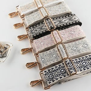 Può includere: Assortimento di tracolle decorative per borse in vari colori, tra cui bianco, nero, rosa e argento, con motivi geometrici. Ogni tracolla ha elementi metallici dorati, tra cui chiusure e anelli rettangolari. Un piccolo piatto contiene chiusure aggiuntive.