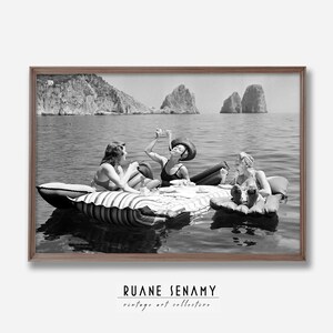 Peut inclure: Trois femmes en maillot de bain se d&eacute;tendent sur un radeau gonflable ray&eacute; dans l'oc&eacute;an. Elles profitent de boissons et de collations tout en flottant dans l'eau. L'arri&egrave;re-plan pr&eacute;sente des falaises rocheuses et un ciel bleu clair.