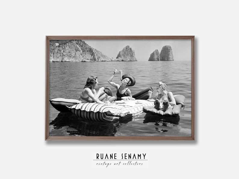 Peut inclure: Trois femmes en maillot de bain se d&eacute;tendent sur un radeau gonflable ray&eacute; dans l'oc&eacute;an. Elles profitent de boissons et de collations tout en flottant dans l'eau. L'arri&egrave;re-plan pr&eacute;sente des falaises rocheuses et un ciel bleu clair.