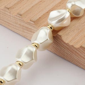 May include: A close-up of a necklace featuring large, irregular, off-white beads. The beads are separated by small gold-colored spacers. The necklace is partially resting on a light-colored wooden surface.