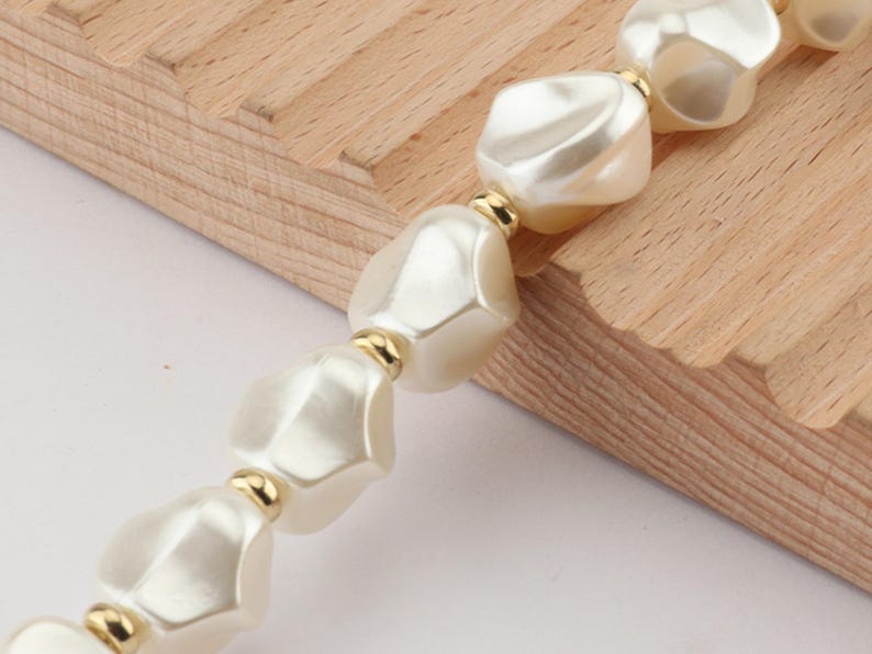 May include: A close-up of a necklace featuring large, irregular, off-white beads. The beads are separated by small gold-colored spacers. The necklace is partially resting on a light-colored wooden surface.