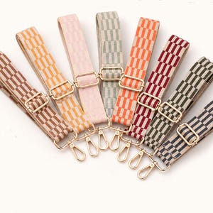 Puede incluir: Una colección de ocho correas ajustables para bolsos dispuestas en forma de abanico. Cada correa presenta un patrón de cuadros tejido en varios colores, incluyendo marrón, amarillo, rosa, verde salvia, naranja, rojo, verde oscuro y azul marino, con herrajes de metal dorado.