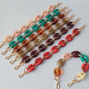 Op de afbeelding: Een collectie van zeven kettingriemen met ovaalvormige acrylschakels in diverse effen en gemarmerde kleuren, waaronder perzik, groen, bruin en rood. Elke riem is aan beide uiteinden afgewerkt met goudkleurige metalen karabijnsluitingen.