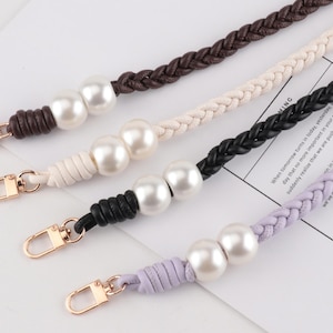 Può includere: Quattro cinturini intrecciati per borse in marrone, crema, nero e lavanda. Ogni cinturino presenta grandi perle bianche e chiusure color oro. I cinturini sono esposti su una superficie bianca.