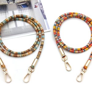 Puede incluir: Dos correas de bolso con cuentas de colores con cierres metálicos dorados. Una correa presenta tonos tierra, mientras que la otra es vibrante con rojo, amarillo y azul. Estas correas están diseñadas para bolsos.