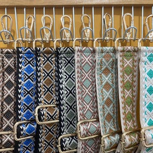 Puede incluir: Nueve correas tejidas de colores con patrones geométricos cuelgan de ganchos dorados. Las correas son en tonos de azul, marrón, verde y rosa.