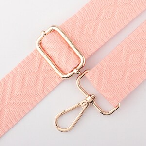 May include: A light pink, patterned bag strap with gold-colored metal hardware. The strap features a repeating geometric design and includes a rectangular buckle and a swivel clasp.