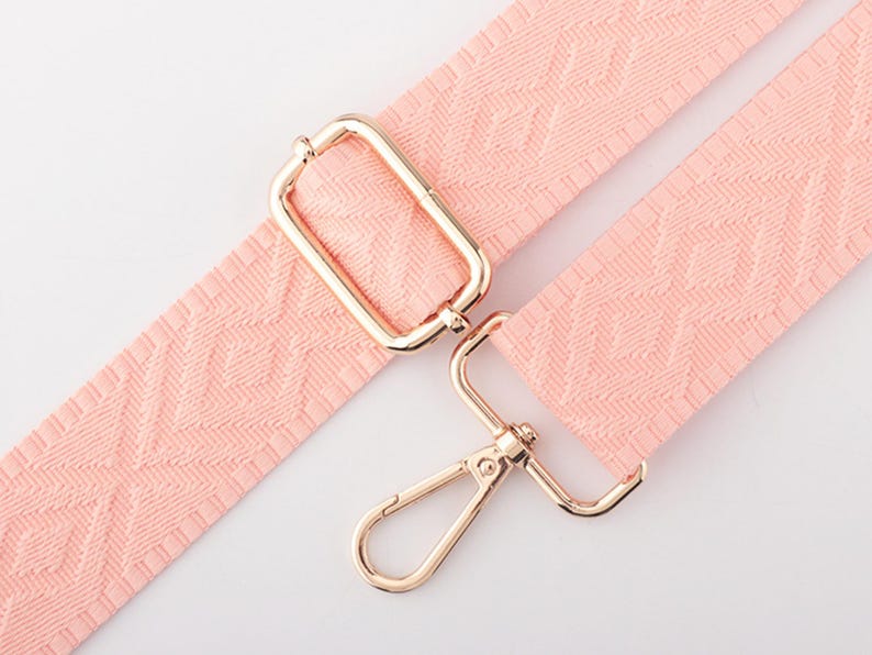 May include: A light pink, patterned bag strap with gold-colored metal hardware. The strap features a repeating geometric design and includes a rectangular buckle and a swivel clasp.
