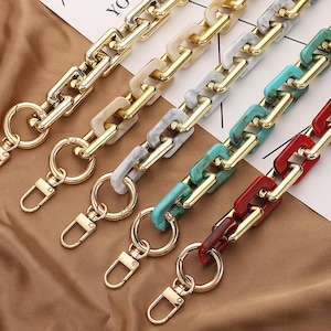 Puede incluir: Primer plano de cinco correas de cadena de bolso coloridas. Cada cadena presenta eslabones de metal dorado y eslabones acrílicos coloridos en tonos crema, gris, turquesa y rojo. Cada cadena tiene un cierre y un anillo dorados.