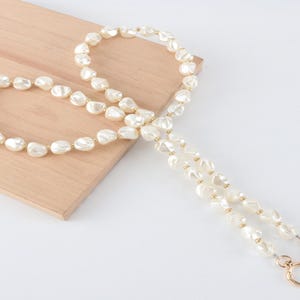 May include: A cream-colored pearl necklace with a gold clasp and accents. The necklace is composed of irregularly shaped pearls and is displayed on a light wooden surface.