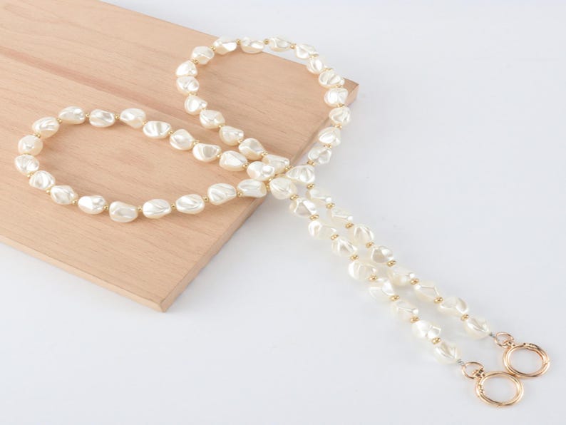 May include: A cream-colored pearl necklace with a gold clasp and accents. The necklace is composed of irregularly shaped pearls and is displayed on a light wooden surface.