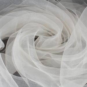 May include: Close-up of white, sheer fabric arranged in swirling patterns. The fabric appears delicate and lightweight, with a soft, flowing texture. The folds and layers create a sense of depth and movement, suitable for bridal or decorative use.
