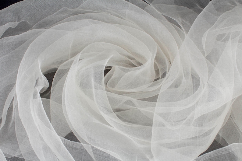 May include: Close-up of white, sheer fabric arranged in swirling patterns. The fabric appears delicate and lightweight, with a soft, flowing texture. The folds and layers create a sense of depth and movement, suitable for bridal or decorative use.