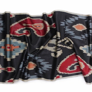 May include: Black fabric with a repeating pattern of red, blue, and cream-coloured shapes. The fabric is draped, showing folds and texture. The design has an ikat style, with a dark background.
