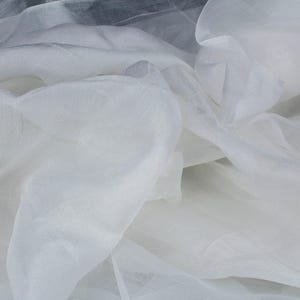 May include: Close-up of white sheer fabric with a delicate, flowing texture. The fabric is layered and creates soft folds and shadows. The material appears lightweight and translucent, suitable for apparel or decorative projects.