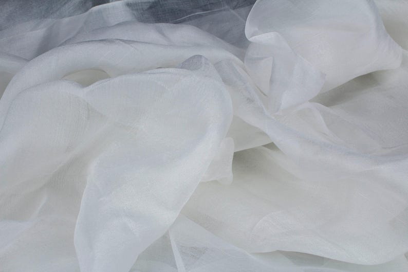 May include: Close-up of white sheer fabric with a delicate, flowing texture. The fabric is layered and creates soft folds and shadows. The material appears lightweight and translucent, suitable for apparel or decorative projects.