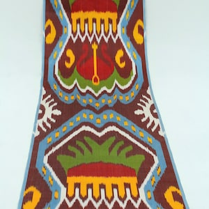 May include: A patterned textile with a repeating design in shades of brown, red, green, yellow, and blue. The design features stylized floral and geometric shapes. The fabric has a soft, flowing drape, suggesting it could be used for clothing or home decor.