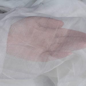 Könnte beinhalten: Nahaufnahme einer Hand unter Lagen aus durchsichtigem, weißem Stoff. Der Stoff wirkt zart und durchscheinend, wodurch der Hautton durchscheint. Die Textur des Stoffes ist sichtbar und erzeugt einen weichen, ätherischen Effekt.