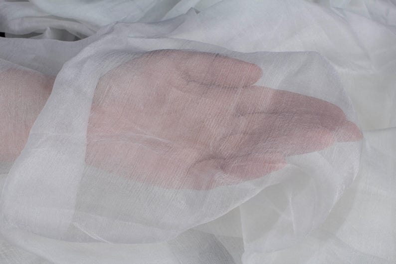 May include: Close-up of a hand beneath layers of sheer, white fabric. The fabric appears delicate and translucent, allowing the skin tone to show through. The texture of the fabric is visible, creating a soft, ethereal effect.