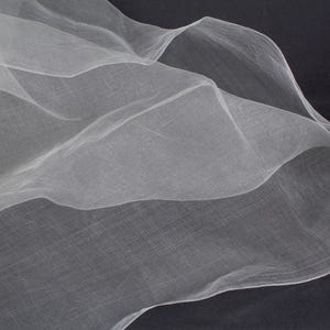 May include: A close-up of flowing, sheer white fabric against a dark background. The fabric has a delicate, airy texture, with soft folds and curves. The image highlights the fabric's translucence and subtle details.