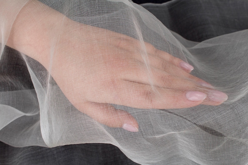 May include: A hand rests on a sheer, white fabric. The fabric is draped over a dark gray surface, creating a contrast in color and texture. The hand has manicured nails.