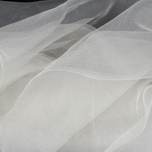 May include: A close-up shot of flowing, sheer white fabric. The fabric is layered and creates soft, undulating waves. The material appears lightweight and delicate, with a subtle texture. The overall effect is ethereal and elegant, suggesting a textile suitable for apparel or decor.