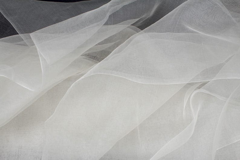 May include: A close-up shot of flowing, sheer white fabric. The fabric is layered and creates soft, undulating waves. The material appears lightweight and delicate, with a subtle texture. The overall effect is ethereal and elegant, suggesting a textile suitable for apparel or decor.