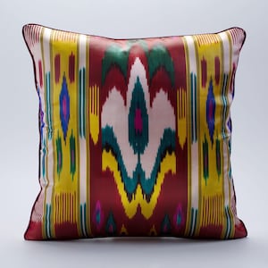 Könnte beinhalten: Dekoratives quadratisches Kissen mit einem lebendigen Ikat-Muster. Das Kissen hat einen bordeauxfarbenen Rand und ein zentrales Design mit weißen, gelben, grünen, blauen und rosa vertikalen Streifen. Auf weißem Hintergrund.