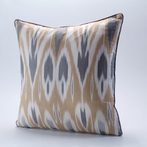 May include: Square cushion featuring an ikat design in beige, grey, and white. The cushion has a dark brown border, suitable for home decoration.