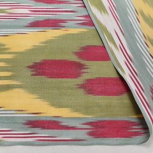 May include: A close-up of a fabric with a colorful ikat pattern. The fabric is a blend of yellow, green, red, and white. The pattern is a traditional ikat design, with a repeating motif of stripes and dots.